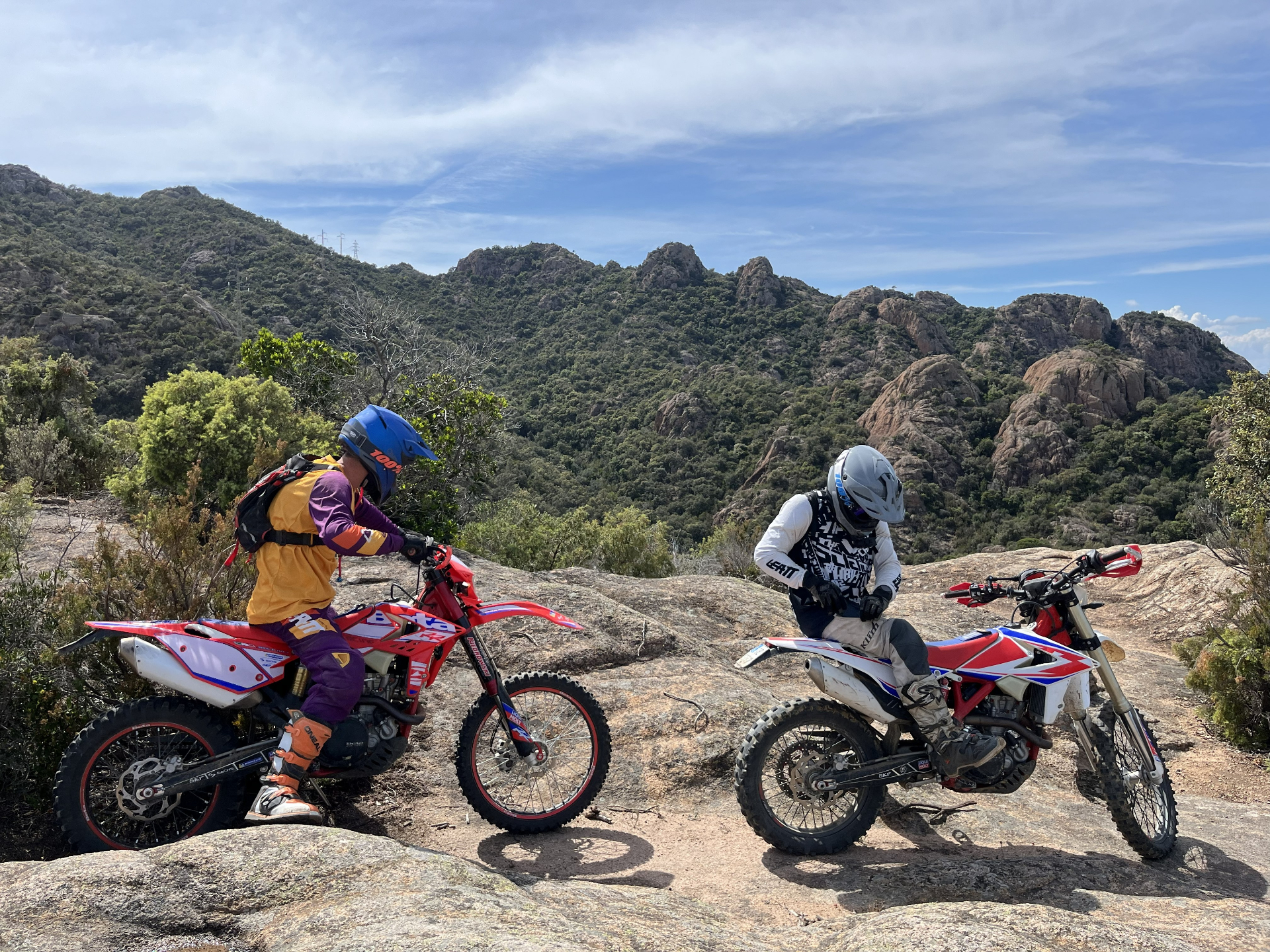 Two enduro riders on rocky terrain with mountain backdrop